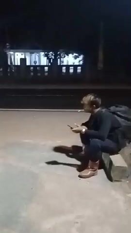 A young person with blonde hair sits on a concrete curb at night by a road. They are wearing a dark long-sleeved shirt, dark pants, and brown boots. A large dark backpack is behind them. The person is holding a smartphone in their left hand and a cigarette in their right hand, with smoke visible. They are looking at the smartphone. In the background, across the road, there is a building with lights on and trees. The area is dimly lit by artificial light.