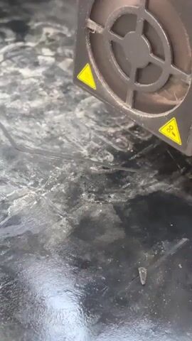 The video shows a close-up of a 3D printer nozzle dispensing black filament onto a dark, textured surface. The nozzle is attached to a black housing with fans and warning symbols. The filament is being laid down in a thin, continuous line, suggesting the beginning of a print. The surface appears dusty and has some circular marks or residue. The lighting is direct and highlights the details of the printing process.