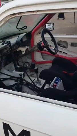 The video shows the interior of a racing car. The car is white with a red roll cage. The seats are black, and there's a steering wheel, gear shift, and electronic dashboard. The back of the car contains a fuel cell and fuel pumps. The words "Meri School" and "Shaw School" are printed on the side of the car. A person in white shorts is standing near the car.