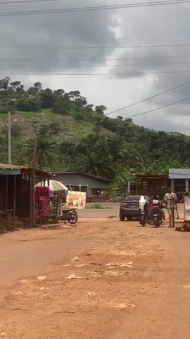 A dirt road passes through a rural settlement with small shops lining the left side. In the background, a lush green hillside rises under a cloudy sky. A few people are walking on the road, and a car is parked nearby. A tricycle-like vehicle is also visible. Signs for "AKAMMIN'S KITCHEN" and "16 16 FARMS FROZEN FOOD DEPOT" are displayed. The scene suggests a typical day in a developing area with local commerce and a natural landscape.