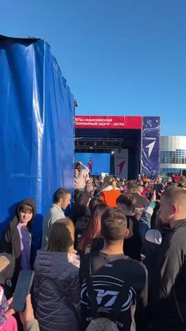 The scene is a crowded outdoor event in front of a stage on a sunny day. The stage has a red and white banner with text in Russian: "ХАНТЫ-МАНСИЙСКИЙ АВТОНОМНЫЙ ОКРУГ - ЮГРА". On the stage, a woman is performing while a video of her singing is shown on a large screen behind her.  The crowd is dense, with people of various ages and attire. Many are holding up smartphones, presumably recording or taking pictures of the performance. A large blue tarp or curtain is visible on the left side, obscuring part of the view. The building behind the stage appears to be modern, with large glass windows.