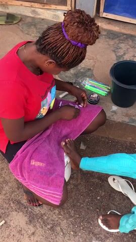 A woman with braided hair, tied in a bun with a purple scrunchie, is squatting outdoors, meticulously performing a pedicure or applying nail decorations to another person's foot. She is wearing a red t-shirt with some visible text and black shorts. A purple towel, embroidered with the word "Elegance," is draped over her lap. The customer's foot, clad in a white flip-flop, rests on a white plastic stool. The customer is wearing light blue pants.

On the ground around the woman are various beauty supplies: a black bucket, a clear container filled with colorful plastic items (possibly beads or nail accessories), and a small black container, potentially nail polish or glue, with text that appears to be "SAIMA" or "MAINA." Several small, light-colored decorative pieces are scattered on the towel and ground.

The scene takes place on a concrete or dirt surface during the daytime, with a building wall and wooden structures visible in the background, indicating an outdoor, casual setting in Enyibichiri, Nigeria.