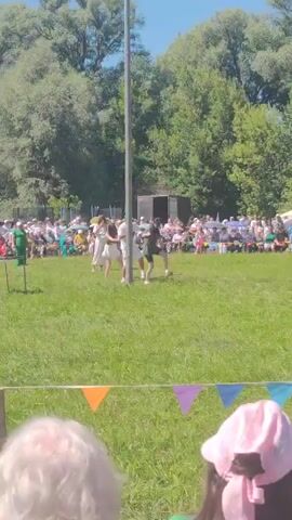 The video shows an outdoor event on a sunny day with a crowd of people watching. In the foreground, the back of a person wearing a pink hat is visible. In the mid-ground, a tall pole stands in a grassy field. Four individuals, three women and one man, are gathered around the base of the pole, appearing to participate in an activity. Two of the women are wearing white dresses, one is wearing a white top and a skirt, and the man is wearing a white shirt and shorts. The crowd is seated in rows behind them, and trees form a backdrop. Decorative flags in orange, purple, and blue are strung across the field in front of the crowd.