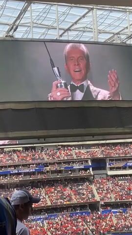 The video shows a large stadium filled with spectators wearing red and white. A large screen at the top displays images and text related to the FIFA World Cup 2026. In the first frame, a man in a tuxedo is shown on the screen holding a trophy. Subsequent frames show the FIFA World Cup 2026 logo and seating section numbers. In the foreground, the backs of two people are visible; one is wearing a white t-shirt and a baseball cap, and the other is wearing an event staff vest. The overall environment is that of a live event or sporting occasion.