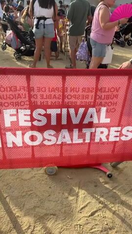The video shows an outdoor festival, possibly a movie screening, with a large gathering of people sitting in chairs on a sandy ground. In the foreground, a red barrier with the white text FESTIVAL NOSALTRES is visible. The barrier has a star-like logo on the left. Behind the barrier, people are milling around and sitting, with some children visible. There are buildings in the background and a large white screen, likely for a projection. The sun is shining, casting shadows. The overall atmosphere appears to be casual and festive.