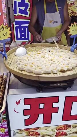 A man in a yellow apron is preparing food at a street stall. He is using chopsticks to place dumplings into a white takeout container. The dumplings are arranged in a large, circular bamboo steamer. The steamer is filled with numerous small, white dumplings with orange filling. In the background, there is a sign with blue Chinese characters. The stall appears to be outdoors or in a covered market.