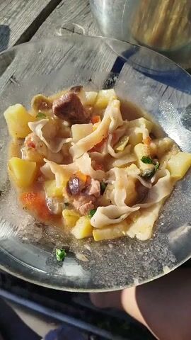 The video shows a close-up, overhead shot of two dishes of food. The dish in the foreground, on a clear glass plate, contains a stew-like meal with chunks of meat, potatoes, and wide noodles in a light brown broth. A spoon is visible at the right edge of the plate. In the background, a stainless steel bowl is filled with a salad composed of chopped tomatoes, cucumbers, onions, and what appears to be a creamy dressing. Both dishes are placed on a wooden surface, likely a picnic table, as evidenced by the wood grain and sunlight filtering through. The lighting suggests an outdoor setting during the day.
