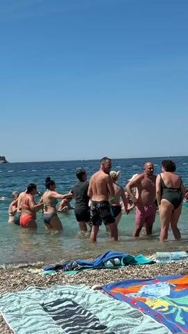 A group of eight people, mostly adults, are standing in shallow water on a sunny day at a beach. The water is clear and calm, with a few buoys visible in the distance. The beach is pebbly, with several towels laid out in the foreground. Four men and four women are present, all wearing swimwear. The group appears to be interacting, with some holding hands and facing each other in a circular formation. Their attire and the setting suggest a casual, recreational atmosphere.