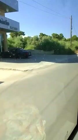 The video shows a view from inside a vehicle looking out at a road. The road is paved and has a white line marking the edge. On the left side of the road, there is a grassy area with trees and bushes. In the background, there is a clear blue sky. At approximately 00:01, a person on a white scooter is seen riding on the road. The scooter is white with a black seat. The person is wearing a white shirt and dark shorts. The rest of the video shows the vehicle continuing to drive along the road.