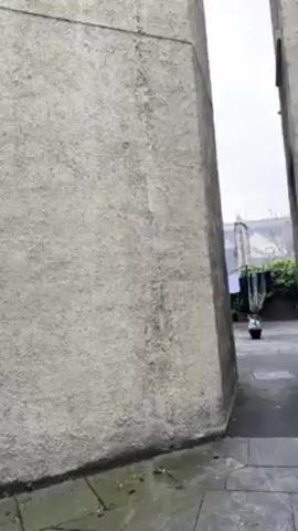 The video shows a slow pan-up and then a panning shot of a courtyard between two buildings on a cloudy day.  The buildings are tall and have a textured gray exterior with evenly spaced white-framed windows. The courtyard is paved with gray square tiles and contains potted plants. A red building is visible in the distance beyond a black iron fence. The person filming walks through the courtyard during the pan.