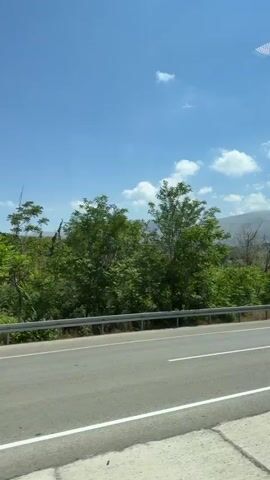 The video shows a view from a moving vehicle. The road is in the foreground, with white lines dividing the lanes. A metal guardrail runs alongside the road. Beyond the guardrail is a landscape with green trees and rolling hills in the background. The sky is blue with scattered white clouds. The scene appears to be rural and possibly mountainous.