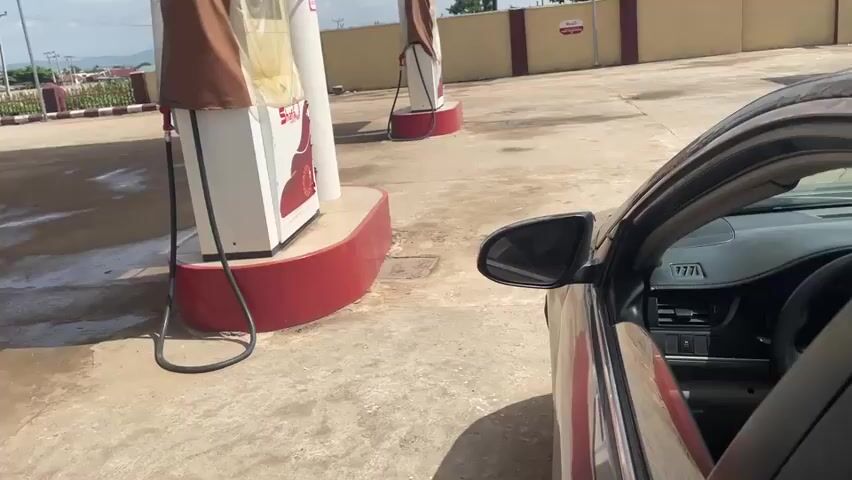 The video begins with a view of a car parked at a gas station. The car's passenger side door is open, revealing the interior. In the foreground, the driver's side of the car is visible, with the steering wheel and dashboard in view. The car is dark in color. The gas station has red and white pumps, with hoses connected to them. The ground is paved concrete, with some wet patches. The weather appears to be bright and sunny, suggesting it is daytime. In the background, there are buildings and vegetation. The scene transitions to an inside view of the car. A person is holding a smartphone, with a video call in progress. The phone screen displays a person on the call. The person holding the phone is wearing a maroon hoodie and a headscarf with a distinctive 'G' pattern. Another person, or part of a person, is visible in the background, covered with a grey fabric. There are bags and other items in the back seat. The overall impression is a moment of waiting at a gas station while engaged in a video call.