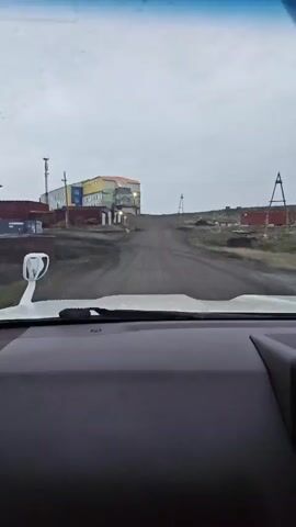 The video shows a view from inside a vehicle, looking out through the windshield. The windshield has water droplets on it. The vehicle is on a dirt road, heading uphill towards a cluster of buildings. The buildings are colorful, with yellow, blue, and red sections, and appear to be low-rise structures. There is a tall antenna or tower to the left of the buildings. The environment appears to be barren and somewhat desolate, with a cloudy or overcast sky. The road is unpaved and dusty. On the right side of the road, there are some scattered rocks or debris and what looks like a small windmill or weather vane. Further in the distance on the right, a piece of heavy machinery, possibly a tractor or bulldozer, is visible. The interior of the vehicle is partially visible in the foreground, including the dashboard and a side mirror. The overall impression is of a remote and possibly industrial or settlement area in a cold or northern climate.