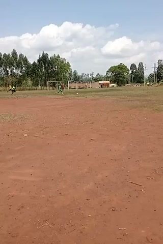 This media file captures an outdoor scene on a sports field under a partly cloudy sky. The ground is a mixture of reddish-brown dirt and patches of dry grass. In the foreground, the dirt area is prominent, with a few scattered blades of grass and some small debris.

The middle ground features a grassy field with a soccer goal visible on the left side. Several individuals are present on the field, engaged in what appears to be a casual soccer game. One person in a green shirt is running with a soccer ball. Another person in a black shirt is also running, potentially participating in the game or observing. A few other figures are scattered across the field, some appearing to be players.

In the background, there are trees, buildings, and utility poles, suggesting a rural or semi-urban setting. The buildings appear to be simple structures, possibly homes or small school buildings. The overall impression is of a community sports activity taking place in a warm, possibly dry, environment, likely during the daytime given the brightness and shadows. The weather appears pleasant with scattered clouds. The image does not contain any visible text. The activity is clearly recreational, with people enjoying a game of soccer.