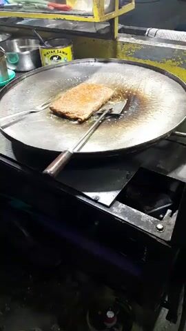 A person's hands are visible, wearing a light-colored sweater sleeve and a darker blue sleeve. The hands are holding two cooking utensils, a spatula and tongs, to manipulate a rectangular-shaped food item on a large, round griddle. The food item appears to be golden brown and is sizzling in oil. The griddle is heated by a black stove. In the background, a yellow structure with containers and a yellow sign with a red rooster illustration are visible. The scene appears to be an outdoor food stall at night.