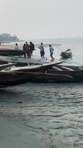 The video shows a group of people near a riverbank filled with wooden boats. Some people stand near a boat that's partially in the water, while others are on or near other boats on the shore. The environment appears to be a river or large body of water with a sandy bank. The weather looks overcast. The video captures a slice of everyday life near the water, presumably focusing on the interaction between people and their boats.