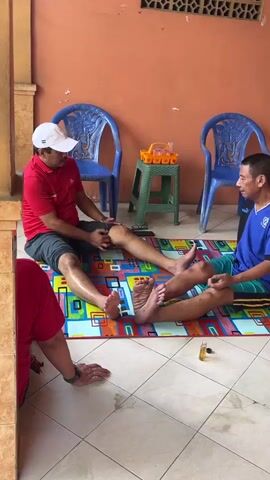 A man with no arms, wearing a red shirt and a white cap, sits cross-legged on a colorful mat. He is facing another man who is wearing a blue shirt and green shorts, and is massaging the first man's legs. The second man is also sitting cross-legged on the mat. There is a small bottle of liquid on the mat between them. In the background, two blue plastic chairs are visible, and a small green stool with an orange container on top. The scene appears to be outdoors, with an orange wall behind the chairs. Another person, wearing a red shirt, is partially visible in the foreground, also sitting on the floor.