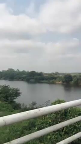 The camera pans across a bridge over a body of water, likely a river or lake. The bridge has a white railing and a roadway with white dividing lines. In the background, there is green vegetation on the banks of the water. The sky is overcast with scattered clouds. Several people walk on the right side of the bridge, and a motorcyclist rides across the bridge. The video ends with a focus on the surface of the bridge, showing some debris on the road.
