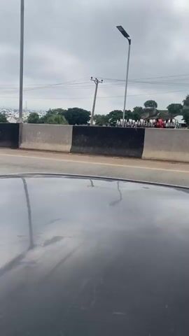 The video captures a view from inside a vehicle looking out towards a highway. In the foreground, the dark, glossy surface of the vehicle's hood is visible, reflecting the overcast sky. A black Toyota Hilux pickup truck is prominently featured in the middle ground, driving on the highway. Further down the highway, a white dump truck loaded with gravel is passing by. In the background, a concrete barrier separates the highway lanes. Beyond the barrier, there are trees, utility poles with wires, and a distant cityscape under a cloudy sky. A group of people are standing on the elevated area beyond the barrier, some are wearing red tops. The overall environment appears to be a busy road during daylight hours with cloudy weather.