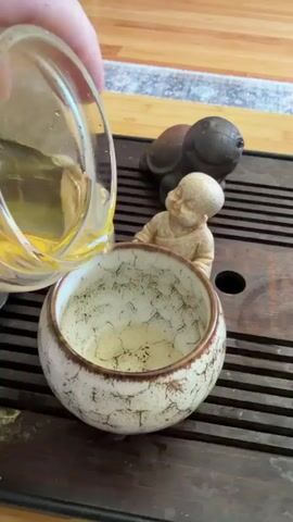A close-up shot shows a person pouring a clear, light-yellow liquid from a glass pitcher into a small, round, ceramic cup. The cup is off-white with brown crackle patterns and holds floating particles of what appears to be tea. The cup sits on a dark-brown, slatted tray. Arranged around the cup are two small, tan-colored Buddha figurines and a dark-gray turtle figurine. The background includes a wooden floor, a rug with gray, blue, and white patterns, and an out-of-focus glimpse of the room. The focus is on the act of pouring the liquid into the cup and the arrangement of figurines around it.