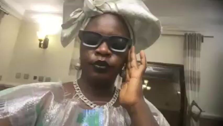 A person wearing sunglasses, a head covering, a silver necklace, and a plastic garment with green, white and blue patterns is visible in a vertically oriented video. A dark belt is seen on the garment. The camera angle shifts and zooms in and out while the person moves. A wall with light fixtures is visible in the background.