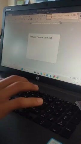A close-up video shows a person using a black HP laptop. The person’s hands are visible as they type on the keyboard. The laptop screen displays a Microsoft Word document with the text "MAVZU: Sanoat tarmoqlari". The laptop is sitting on a wooden table. In the background, there is a blurred view of what appears to be a classroom or a similar space with desks and chairs. The lighting is moderate, and the camera angle is slightly angled downward toward the laptop and hands.