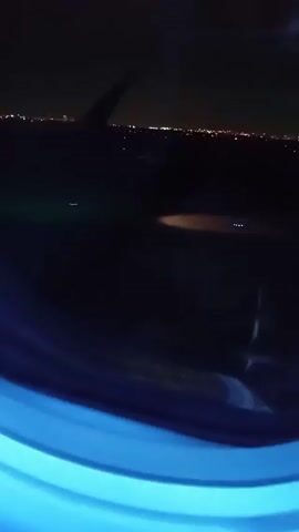 The video captures a nighttime view from an airplane window. The primary focus is the dark outside environment, illuminated by various lights.

In the initial frames (0:00-0:01), a portion of a bright red airplane wing is visible in the upper left section of the frame, extending diagonally downwards. Below the wing, the dark ground or tarmac is visible, with several small, bright blue-ish lights scattered across it. In the far distance, a horizontal line of blurred, warm-toned city lights creates a subtle glow against the dark horizon. The bottom of the video frame is dominated by the airplane window's interior casing, which glows with a vibrant, artificial blue light, creating a stark contrast with the darkness outside.

As the video progresses (0:01-0:03), the red airplane wing gradually moves out of the frame, disappearing by 0:02. The view shifts slightly, indicating the airplane is in motion, likely taxiing on an airport runway or tarmac. The blue lights on the ground appear to move past the window, and a larger, elongated light spot (possibly a reflection or a ground light) briefly appears in the lower-middle section of the external view before fading. The distant city lights remain a consistent presence, twinkling softly.

Throughout the video, the blue glow of the window frame is constant, highlighting the circular shape of the window. The overall scene conveys the atmosphere of a night flight, with the contrast between the plane's interior light and the vast darkness outside, punctuated by man-made lights. There are no people visible, and no text is present in the video.