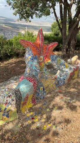 A mosaic-covered bench is in the foreground of the video. The bench is shaped like a creature with a starfish-like head and a body that curves. The mosaic is made of small, colorful tiles in various shades of blue, green, red, yellow, and white. The bench is situated outdoors on a dirt path, with dry leaves scattered around. Behind the bench, there is lush green vegetation, including bushes and trees. A large tree with a thick trunk is to the right of the bench. In the background, there is a panoramic view of a valley with a town or city spread out below. Hills and mountains are visible in the distance under a partly cloudy sky. The sun is shining, casting shadows on the ground. The camera slowly pans to the right, revealing more of the landscape and the town in the valley.