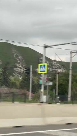 The video shows a view from a moving vehicle, possibly a car, traveling on a highway or major road. On the right side of the road there is a concrete barrier. In the distance, there is a mountain range or a large hill covered in green vegetation. Some areas of the mountain appear to have exposed white rock or possibly construction/mining activity. A line of tall, electrical poles with wires runs alongside the road, partially obscuring the view of the mountain. There are trees and some buildings visible between the road and the mountain. The sky is overcast, and the overall lighting is dim. Several cars are visible on the other side of the road, moving in the opposite direction. Street lights are periodically seen, extending overhead.