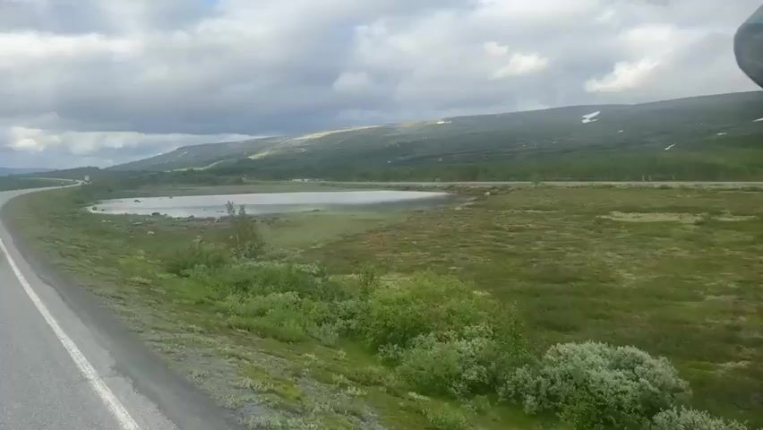 This video captures a scenic drive through a mountainous landscape, likely in Røkland, Norway. The main subject is a paved road, curving to the left and stretching into the distance. Beside the road, a tranquil lake reflects the sky, and on the other side, rolling green hills rise to meet a dramatic cloudy sky. The time of day appears to be late afternoon, with the sun casting long shadows across the hills. The weather is mixed, with patches of blue sky peeking through heavy, dramatic clouds. There are no people or specific objects visible, other than the road, the lake, and the natural elements of the landscape. The overall mood is one of peaceful travel through a remote and beautiful natural setting. The terrain suggests a northern climate, consistent with the provided location context.