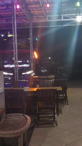 The video pans across an outdoor seating area at night. In the foreground, there are wooden tables and chairs. Beyond the seating area, there is a stage or performance area with lights and people. There are also large metal food serving containers visible. The overall atmosphere appears to be that of an outdoor event or restaurant.