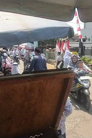 The video captures an outdoor scene, likely a school or community gathering. In the foreground, a wooden object, possibly a table or a cart, occupies the lower right quadrant. Beyond this, a paved surface is visible, with several individuals walking or standing. Two people are seated on a motorcycle. One person is wearing a maroon hijab and maroon dress, and the other is wearing a pink hijab and a maroon dress. A man in a blue shirt and a black skullcap is visible in the middle ground, bending over. Several other individuals are seen in the background, some wearing white and dark attire, suggesting uniforms. Red and white banners or flags are displayed, and a green banner is also present. The environment appears to be sunny, with shadows cast on the ground. The overall impression is of a lively, possibly festive, event.