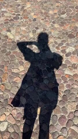 The video shows a shadow of a person on a cobblestone ground. The shadow is of a person wearing a dress. The person is moving their arms. The person is likely taking a video or a picture of themselves. The ground is made of small, multi-colored stones. The lighting suggests it is daytime and the sun is out.