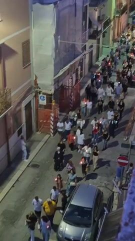 A high-angle shot captures a procession of people walking down a narrow, paved street at dusk. The street is lined with buildings, some of which are under renovation, indicated by scaffolding and orange construction barriers. Most of the individuals in the procession are holding small, lit candles or torches, casting a warm glow. They are diverse in age and attire, with men, women, and children participating. Some are walking in groups, while others are more spread out. Cars are parked along the side of the street, and a few are visible in the distance. A red car is parked further up the street, and a silver car is closer to the foreground. Traffic signs, including a red circle with a diagonal bar, are visible. The overall atmosphere suggests a religious or celebratory event taking place in a residential urban setting. The sky is dark, indicating the late hour.