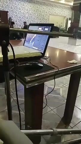 The video shows a desk with a laptop and a mouse. On the laptop screen, there is a program with what appears to be editing software or a simulation. In the background, there are people walking and sitting, and the environment looks like an indoor space with patterned wallpaper and tiled flooring. There is a microphone stand in the foreground.