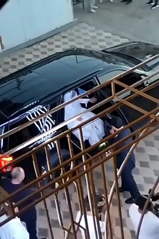 A bride in a white dress and veil is being helped out of the rear passenger side of a black SUV by two men in dark suits. The men are assisting her by holding her arms and guiding her. The bride appears to be holding a bouquet of flowers. The vehicle is parked on a paved area, possibly a driveway or courtyard, with a decorative metal railing in the foreground. There are other people visible in the background, some standing and some in motion, and a camera operator is positioned in the lower right of the frame. The lighting suggests it is daytime.
