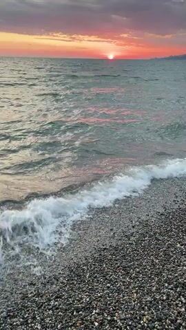 A video clip of a sunset over the ocean. The sun is setting over the horizon, casting a warm glow across the sky. The water is choppy, with small waves crashing on the shore. The beach is covered in pebbles.