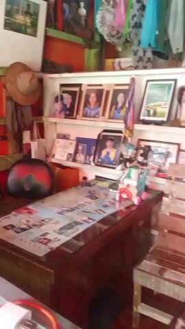 The video pans across a room that appears to be a combined living space and small shop. On the left, a desk is covered with papers and various items, including what appear to be photographs in frames. A bookshelf behind the desk holds more framed pictures, awards, and decorative items. To the right of the desk is a wooden chair with a clear plastic covering. Further to the right, a blue sofa or bed with a star pattern is visible, with clothes and other items on top. A person, wearing a blue and white striped shirt and white shorts, is standing to the right of the sofa, holding a yellow garment. The room has orange walls and tiled flooring. In the background, shelves hold various items, and a television is mounted above a glass display case. A fan is also visible to the right.