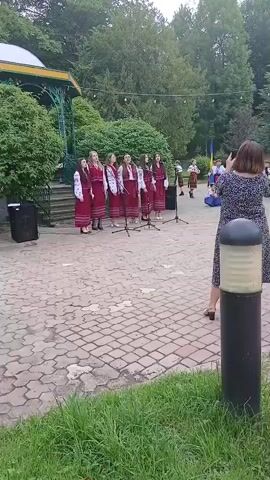 A group of eight young women wearing traditional red and white embroidered dresses are standing in a line and singing outdoors. They are positioned in front of a bandstand with a green roof, surrounded by trees and a paved walkway. A woman in a dark dress is standing in the foreground, to the right, holding up a phone to record the performance. There are speakers set up on either side of the singers. The ground is paved with interlocking bricks and there is green grass in the lower portion of the frame.