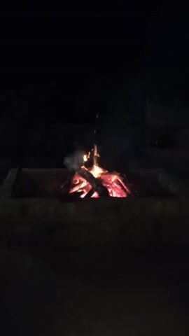 The video shows a campfire burning in a stone fire pit at night. The fire consists of several logs arranged in a crisscross pattern, with flames flickering upwards and smoke rising into the darkness. The embers glow red and orange, illuminating the pit. The surrounding environment is dark.