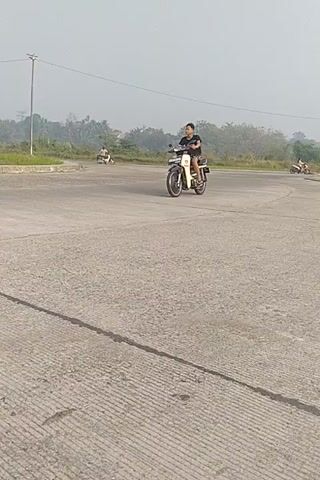 A person is riding a motorcycle on a paved road. The person is wearing a dark t-shirt and shorts. The motorcycle is light-colored. The road is empty except for the motorcycle and rider. There are some buildings and trees in the background. The sky is overcast.
