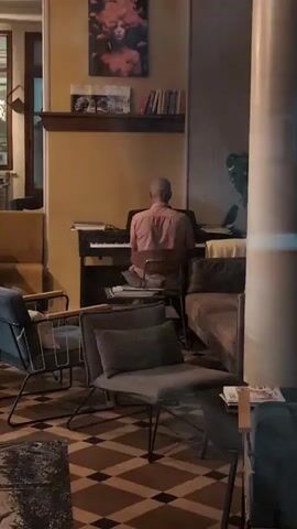 A person is playing the piano in a dimly lit room. The person is seen from behind, and is wearing a pink shirt. The piano is an upright piano. There are several chairs and a couch in the room. There are also books on a shelf above the piano and a painting on the wall. The floor has a patterned tile design.