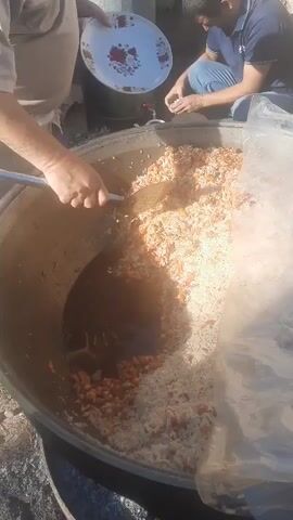 The video shows a close-up view of a large pot filled with rice and what appears to be meat or vegetables. Two people are visible. One person, wearing a light-colored shirt and apron, is using a large metal ladle to stir the contents of the pot and place some of the food onto a white plate with a floral pattern. Another person, wearing a blue shirt and jeans, is kneeling and appears to be handling small white objects. The environment appears to be outdoors, possibly a gathering or event, with natural lighting. A clear plastic sheet is visible draped over the side of the large pot.