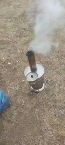 A silver samovar is outdoors with smoke coming from its chimney. The ground is covered in dry grass and dirt. There is a blue bag on the left side of the frame.