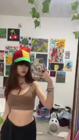 A young woman is standing in front of a mirror, recording a video of herself with her phone. She is wearing a brown crop top, black pants, a colorful hat, and several pieces of jewelry on her wrists and fingers. The woman has dark hair and bangs. She poses for the camera, smiles, and makes a peace sign with her fingers. Behind her is a white wall adorned with various paintings and artwork. There are also decorations hanging from the top of the wall, and a small heater on the floor. A bed is partially visible to the left of the woman.