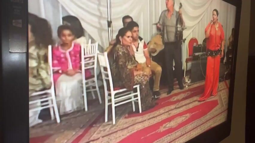 The video shows a scene being played out on a television screen. A band and a belly dancer are performing at what appears to be a wedding celebration. Several people are sitting on white chairs. The dancer is wearing a red outfit and a decorative belt. The band has a man playing a string instrument, another with a guitar, and a female singer. The event is indoors with white draped backdrops. The angle of the camera shows the television from a slight side perspective.