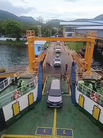 The video captures a view from the deck of a ferry. In the foreground, the green deck of the ferry is visible, with yellow lines marking lanes. A silver car is parked on the ferry. Further ahead, a ramp connects the ferry to a dock. Several cars and people are on the dock. The dock is lined with yellow structures and leads to a terminal building. Mountains and a body of water are visible in the background. The sky appears overcast. The overall scene depicts a ferry terminal with vehicles and passengers preparing for or disembarking from a ferry.