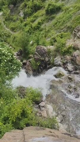 The video shows a cascading waterfall flowing over rocks in a lush, green mountainous environment. The camera angle is high, looking down at the water as it tumbles through a rocky ravine surrounded by vegetation. The water appears white and frothy, indicating a fast flow. Large rocks and greenery are visible on the sides of the waterfall, with a steep, grassy slope in the background. The scene is natural and appears to be in a wilderness area. There are no people or artificial objects visible in the video.