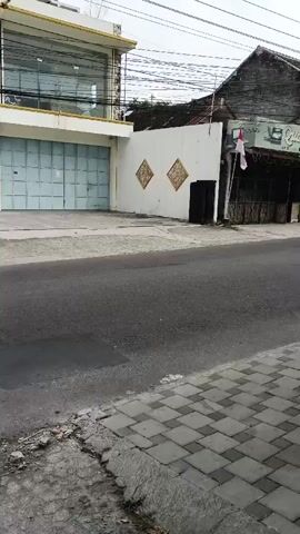 A black scooter with a rider wearing a helmet and a dark shirt turns right onto a paved road. The scooter passes a building with a large, light-colored metal door and a white facade with decorative diamond patterns. Further down the road, a black pickup truck with a cage on the back is seen driving away. The truck is carrying several cardboard boxes. The surrounding environment consists of buildings with tiled roofs, a white fence, and power lines overhead. The road surface is asphalt with some patches of debris and water.