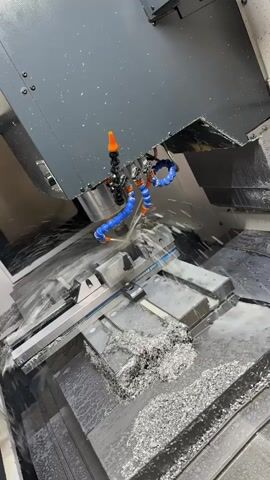 The video shows a close-up view of a CNC milling machine in operation. The machine is actively cutting a piece of metal, creating a significant amount of metallic shavings. A coolant or lubrication fluid is being sprayed onto the cutting area to cool the workpiece and tool, and to help remove the chips. The workpiece is held securely in a vise on the machine's table. The environment appears to be a workshop or manufacturing facility, with the machine occupying most of the frame. There are no people visible in the video. The lighting is somewhat dim, but sufficient to illuminate the active cutting process.